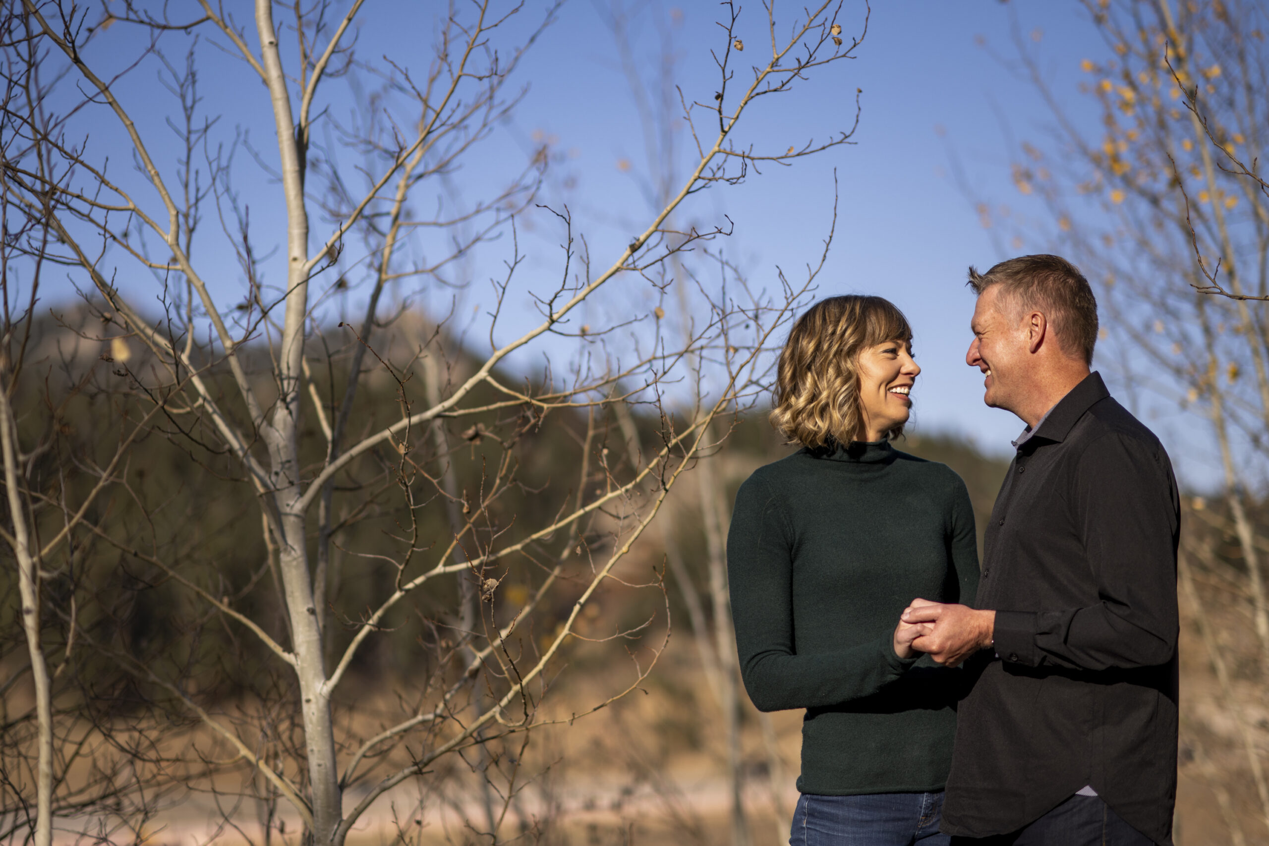 A smiling woman in a green turtleneck faces a man in a black-collared shirt smiling and surrounded by trees during an engagement session at Meyer Ranch Park in Colorado.