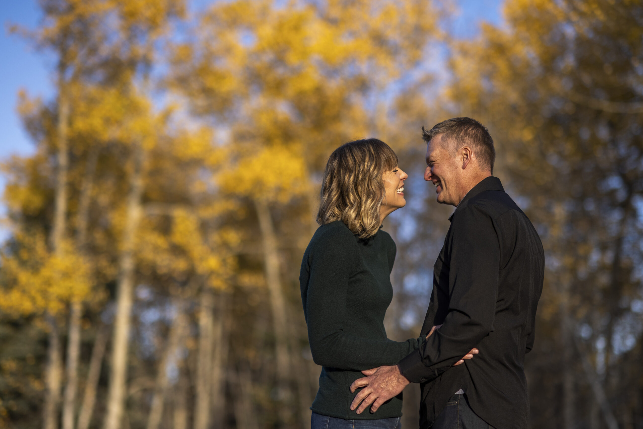 A woman in a green shirt smiles facing a man in a black shirt during an engagement session at Meyer Ranch Park in Colorado.