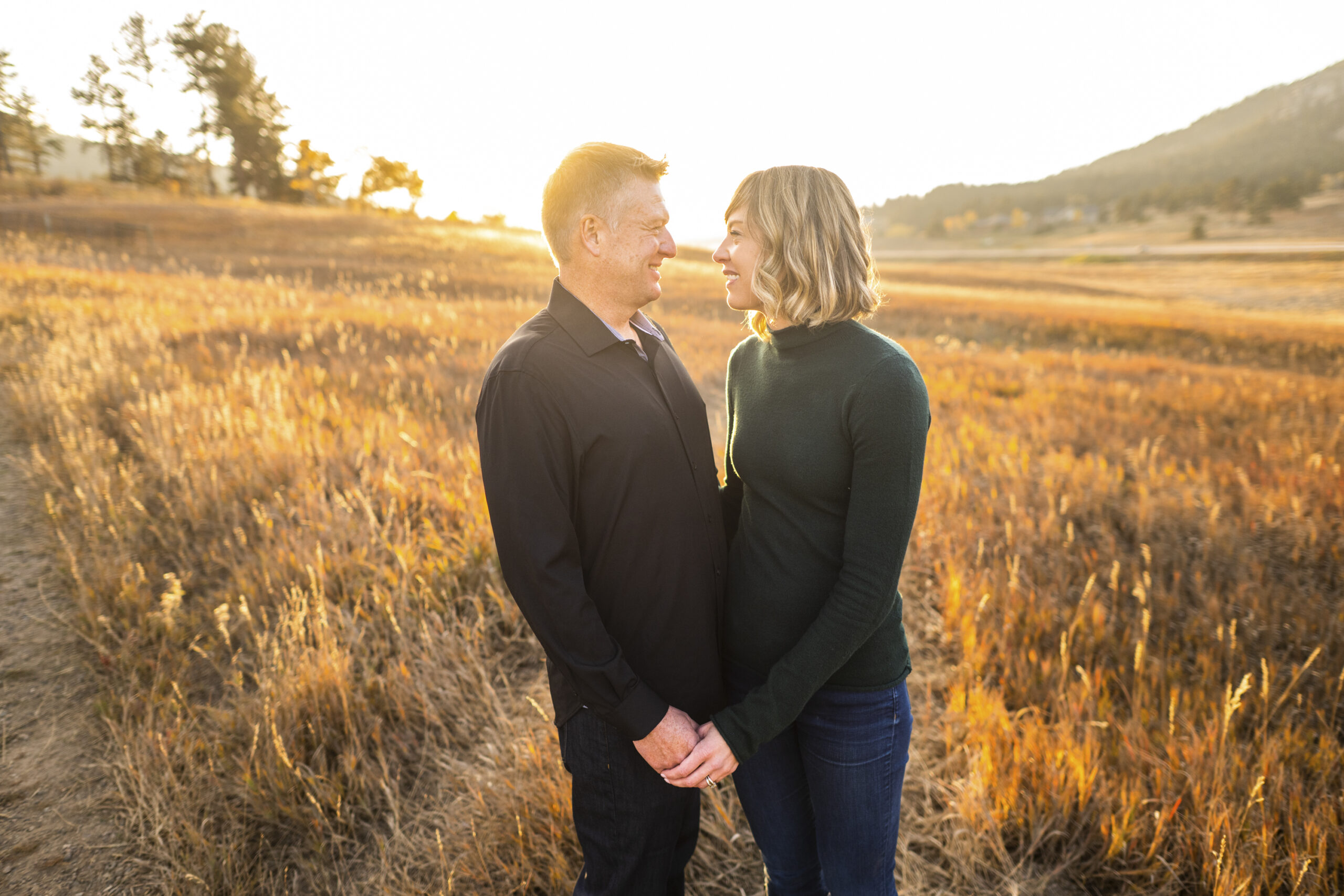 A man in a black-collared shirt and a woman in a green turtleneck look at each other while holding hands with the sun setting in the background during an engagement session at Meyer Ranch Park in Colorado.