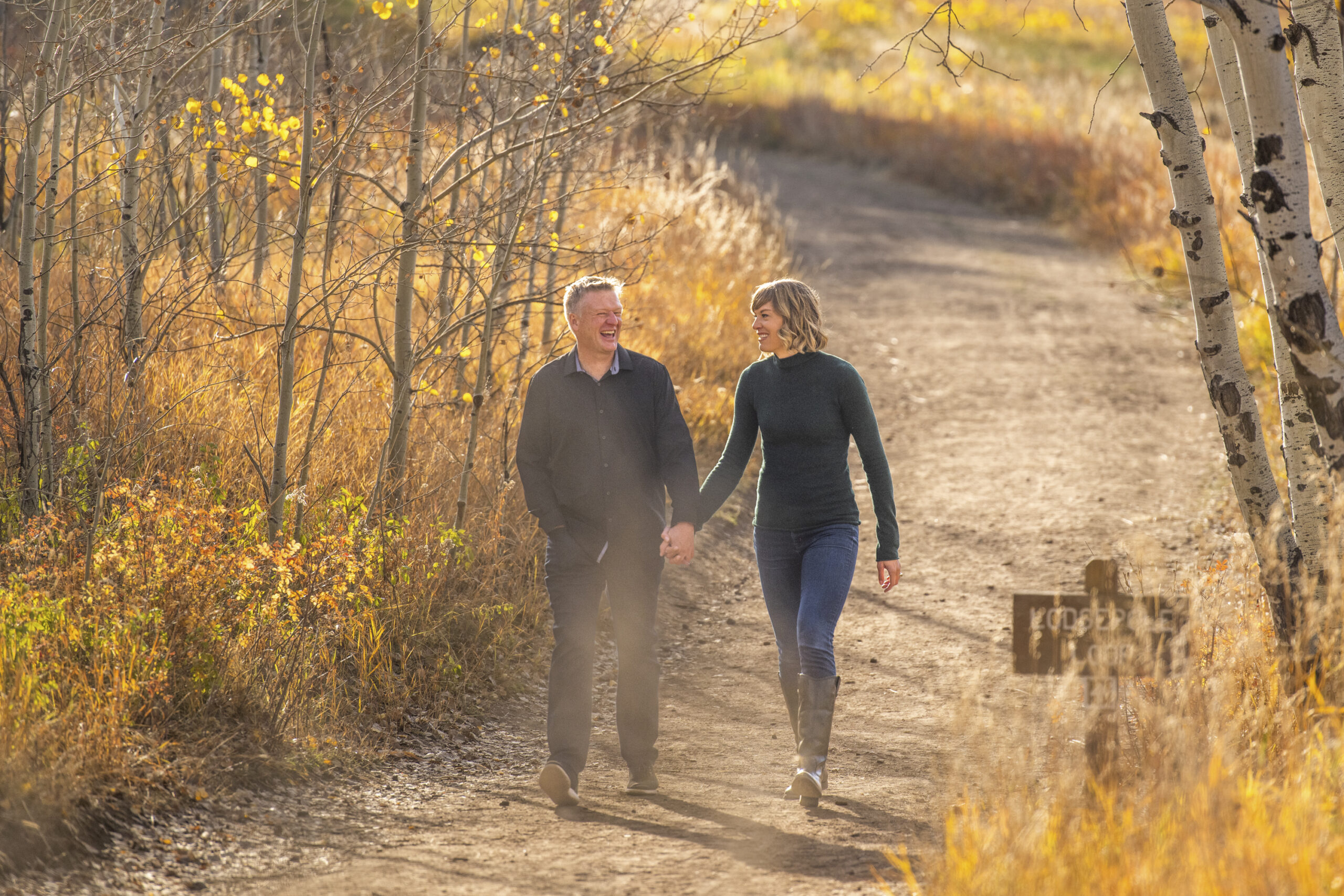 A man in a black-collared shirt and a woman in a green turtleneck walk holding hands on a dirt path during an engagement session at Meyer Ranch Park in Colorado.