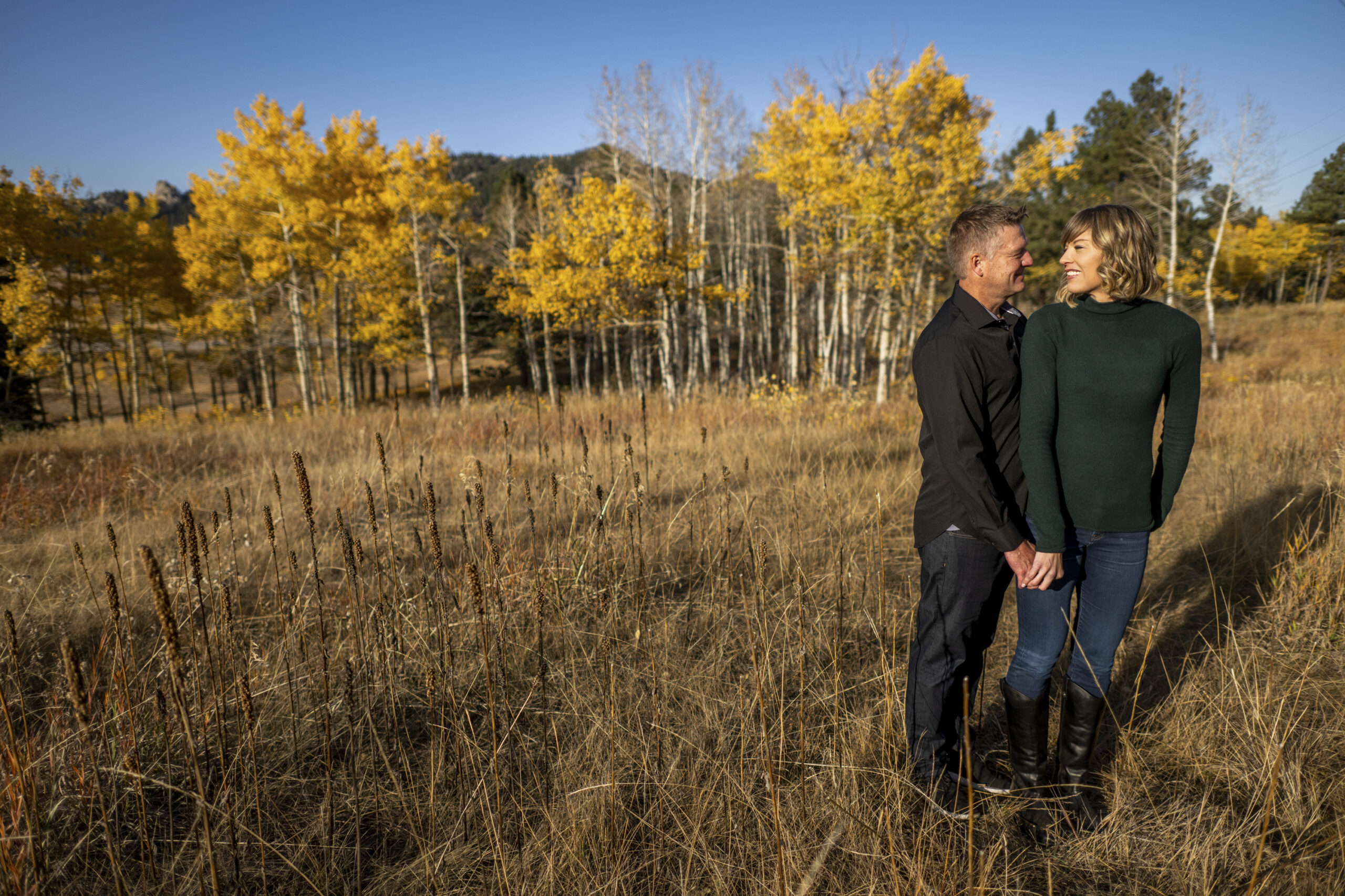 A woman in a green turtle neck stands in front of a man in a black-collared shirt and looks back at him in a field surrounded by trees with yellow trees during an engagement session at Meyer Ranch Park in Colorado.