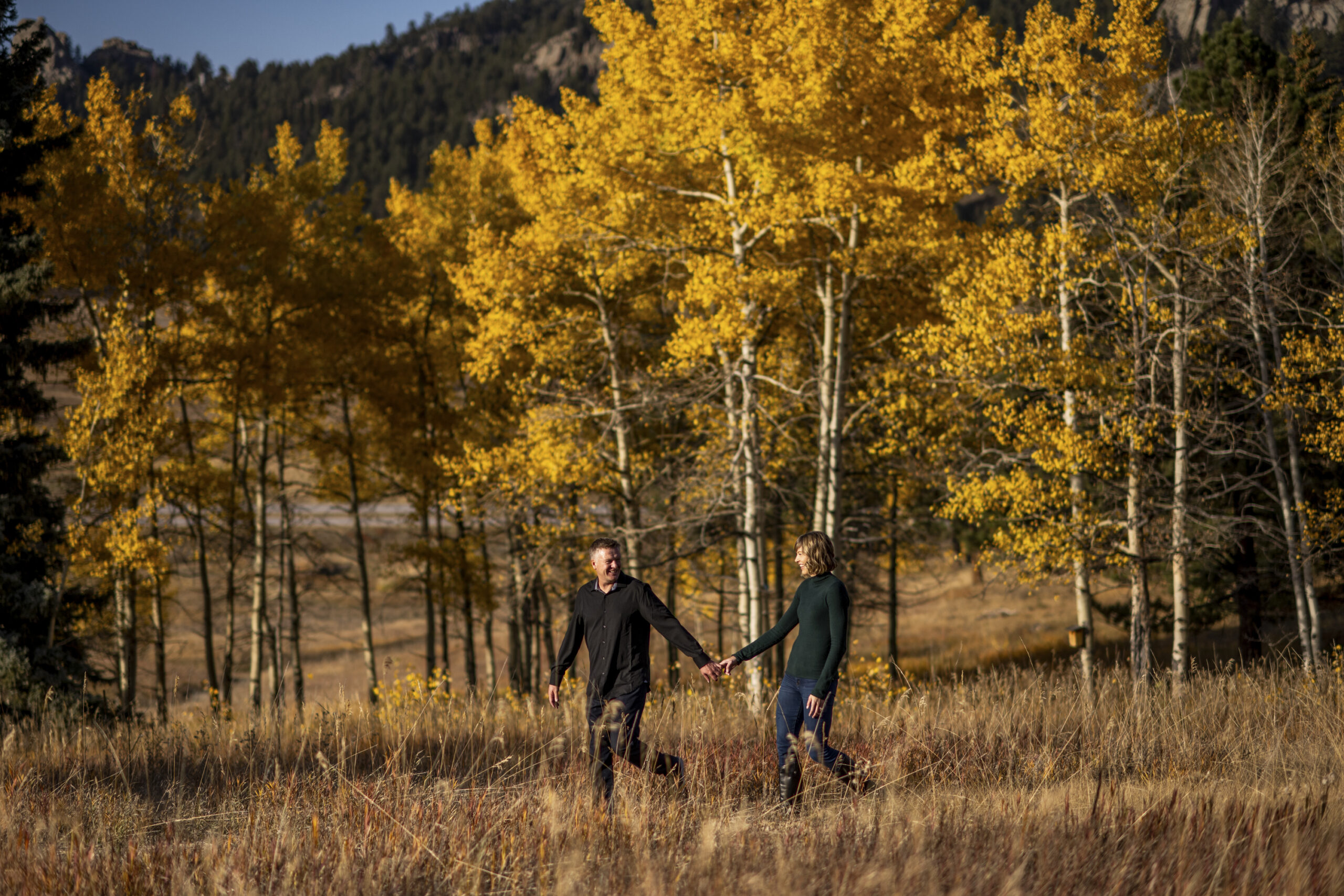 A man in a black shirt leads a woman in a green shirt during an engagement session at Meyer Ranch Park with yellow fall leaves in the background.