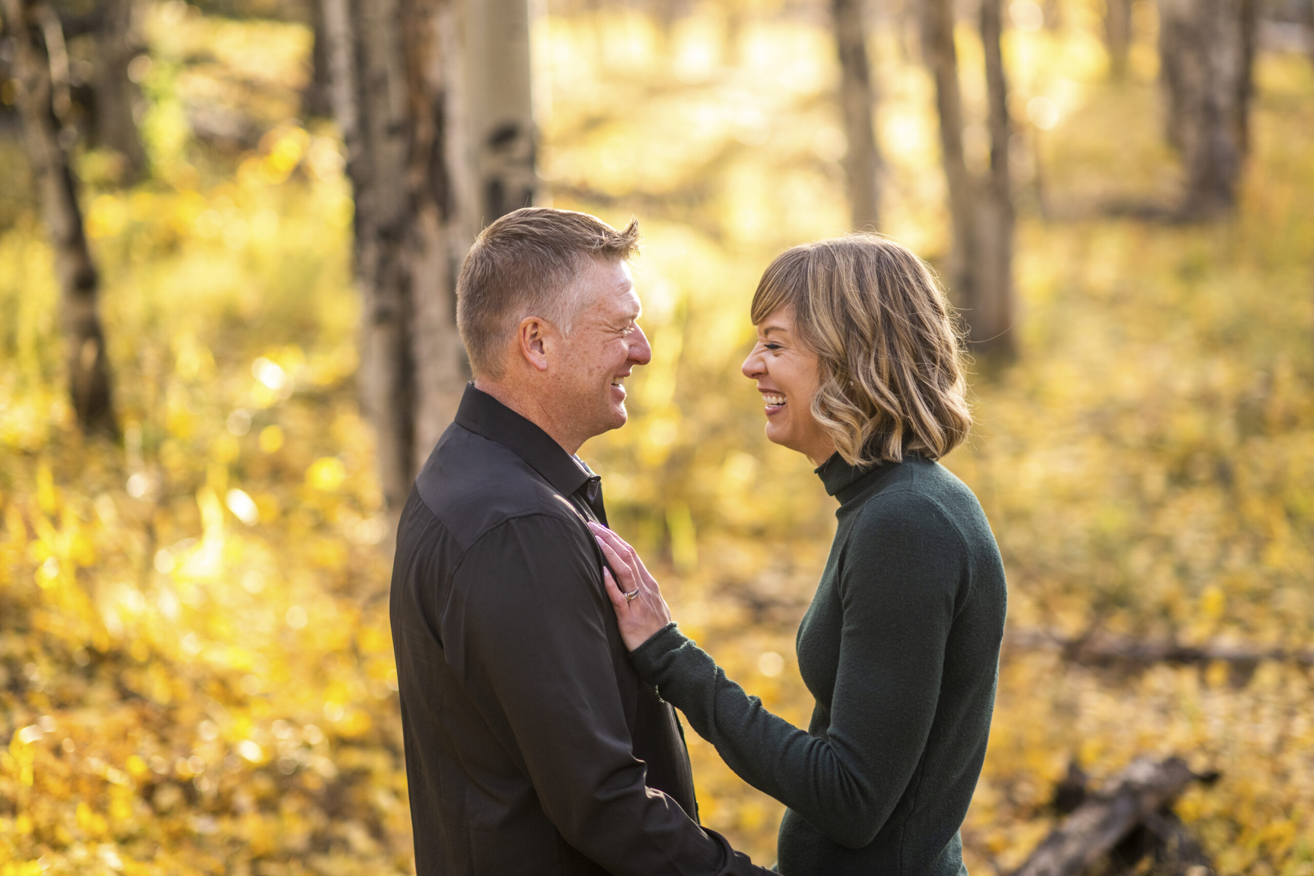 A woman in a green shirt smiles with her hand on a man in a black shirt surrounded by yellow fall colors during an engagement session at Meyer Ranch Park in Colorado.