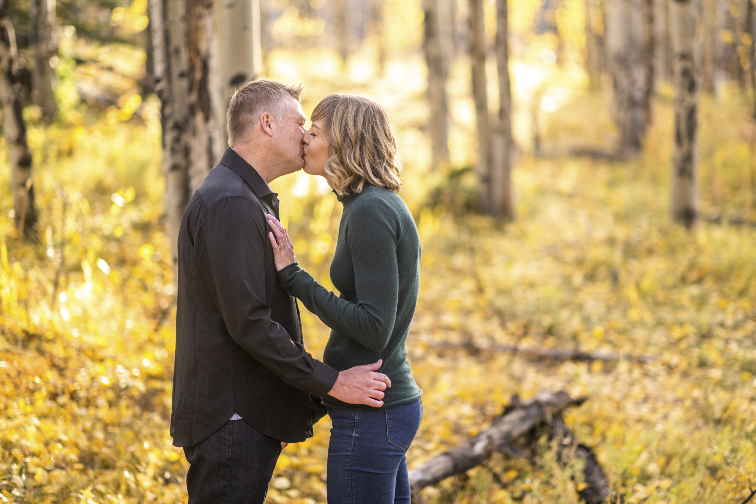 A woman in a green shirt and jeans kisses a man in a black shirt and jeans surrounded by yellow leaves during an engagement session at Meyer Ranch Park in Colorado.