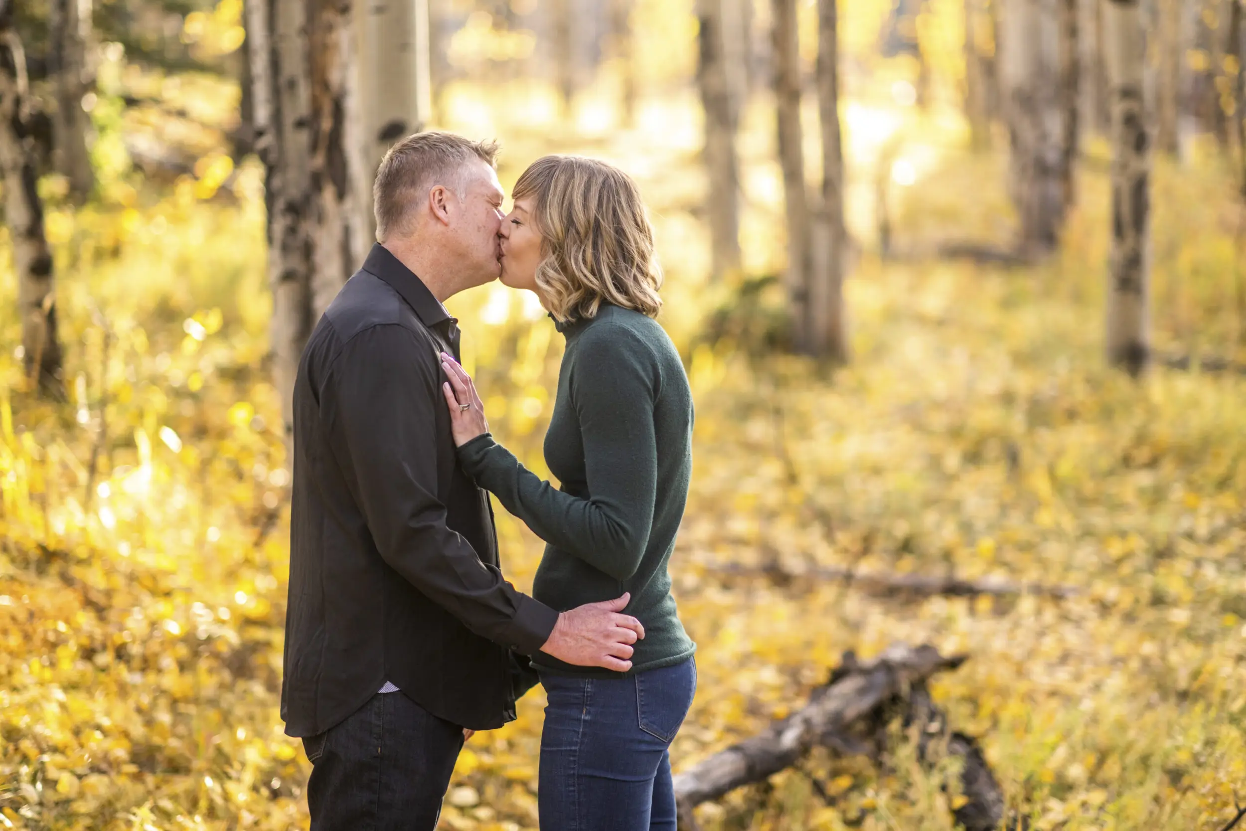 A woman in a green shirt and jeans kisses a man in a black shirt and jeans surrounded by yellow leaves during an engagement session at Meyer Ranch Park in Colorado.