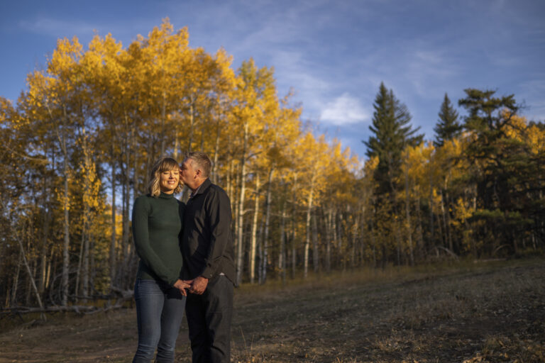 A man in black shirt kisses a woman in a green shirt surrounded by tall trees with yellow leaves in the fall during an engagement session at Meyer Ranch Park in Colorado.