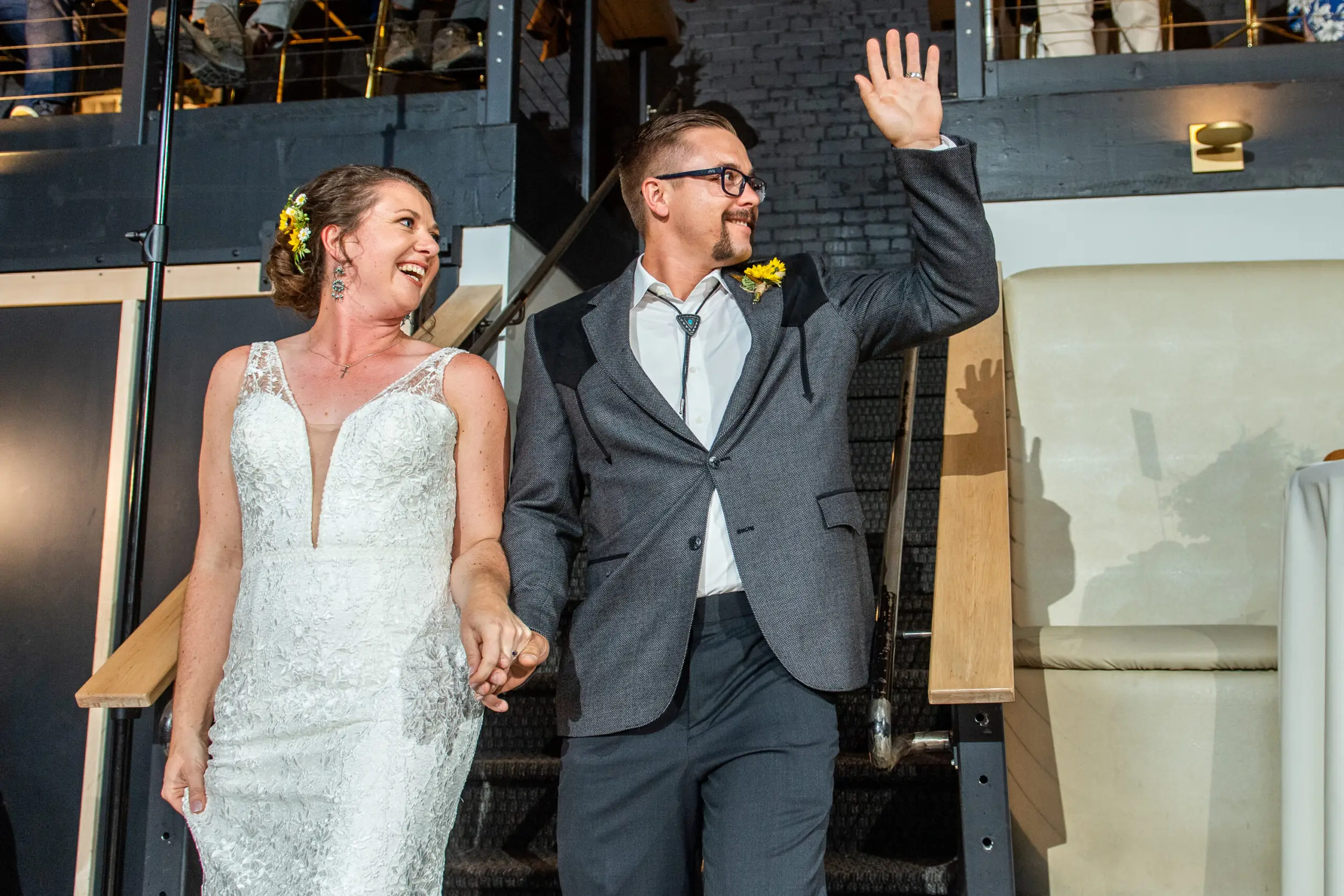 A groom waves while holding his bride's hand as they process in for their wedding reception at the Rose Event Center in Golden, Colorado.