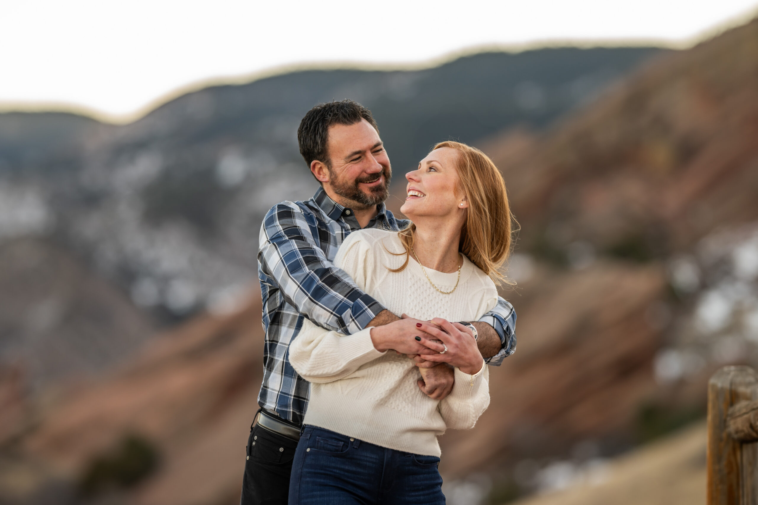 A man wraps his arms around his fiance and they smile at Red Rocks Park and Amphitheatre during a Red Rocks engagement session in Morrison, Colorado.