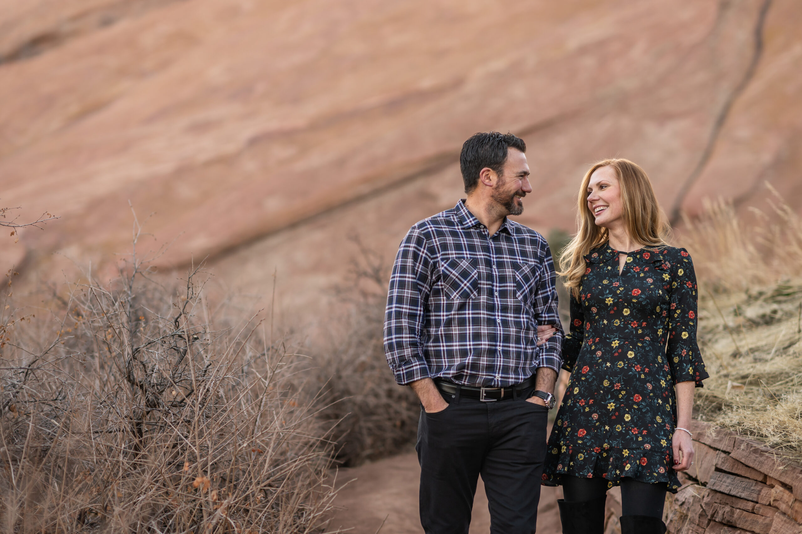 A couple holds hands and laughs while walking along a trail at Red Rocks Park and Amphitheatre during a Red Rocks engagement session in Morrison, Colorado.