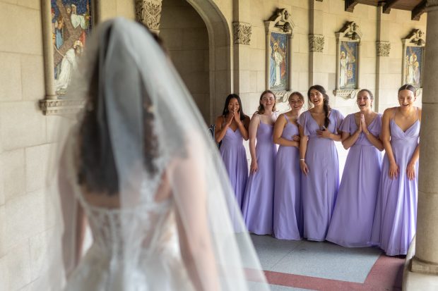 A bride reveals herself to her bridesmaids, wearing purple dresses, for the first time in her wedding dress.