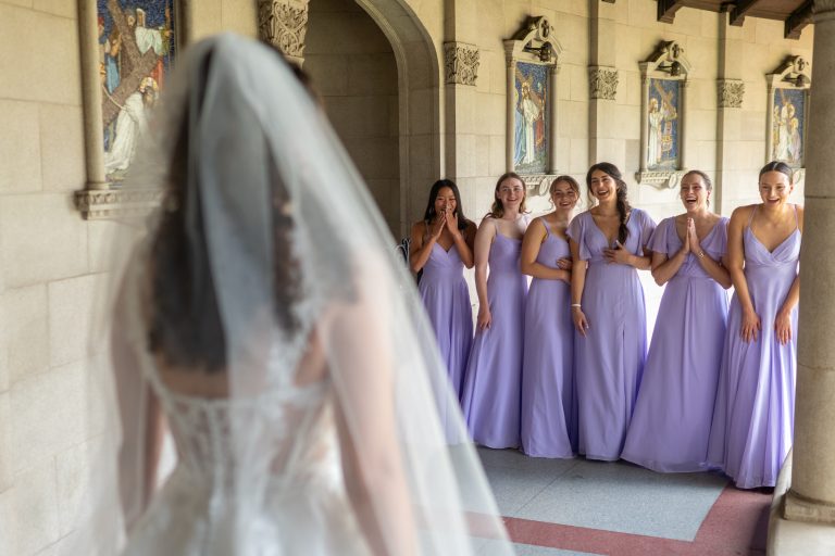 A bride reveals herself to her bridesmaids, wearing purple dresses, for the first time in her wedding dress.