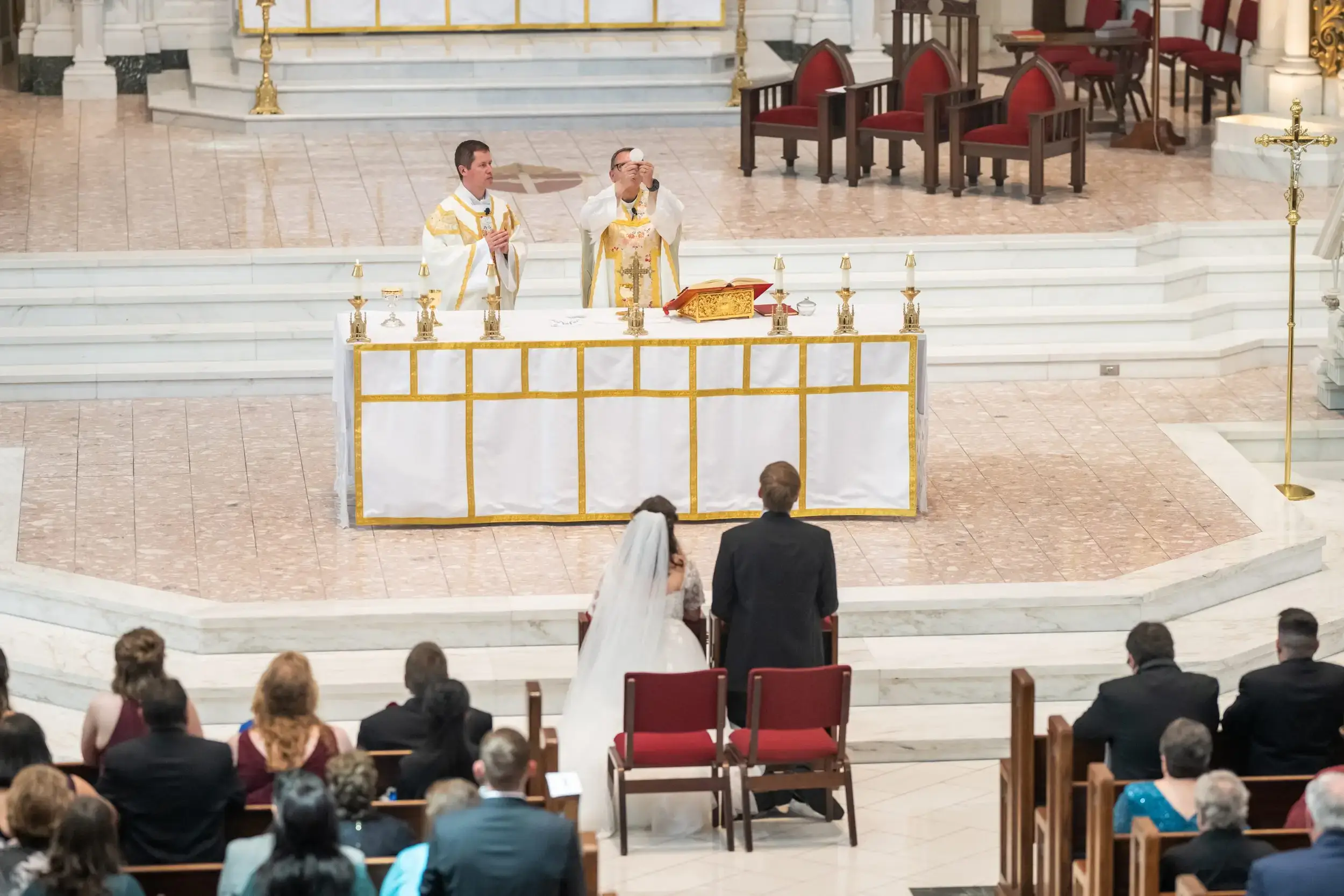A priest consecrates the Eucharist as a bride and groom kneel facing the altar during a nuptial wedding Mass.