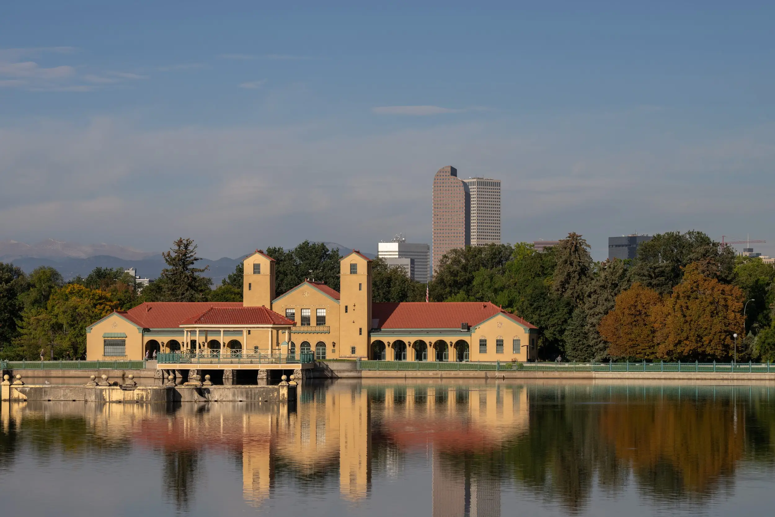 The boat house at City Park in Denver is seen against the city's skyline and mountains in the warm orange morning light.